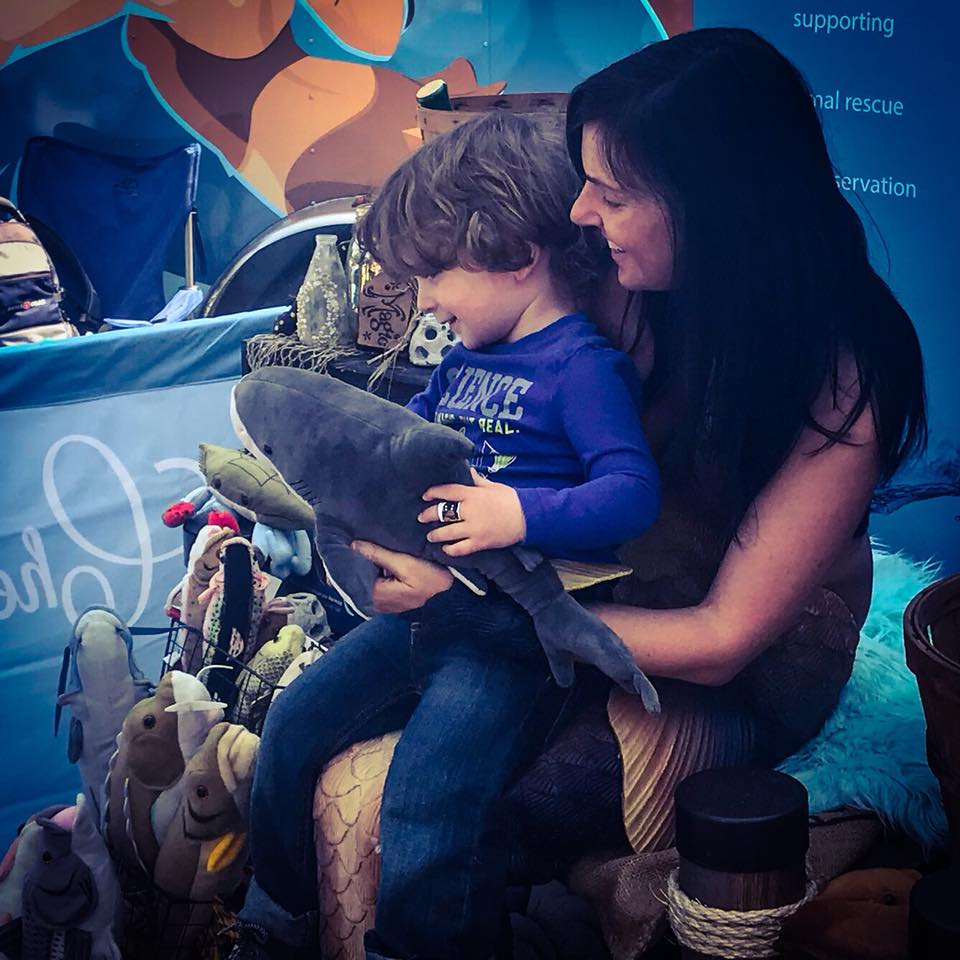 A child holding a plush shark sitting on the Chesapeake Mermaid's lap